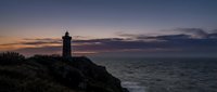 Lighthouse silhouette on rocky cliff overlooking ocean at twilight with dramatic purple and orange sunset sky creating peaceful coastal seascape.