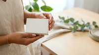 Young woman with medium skin tone holding blank white business card in hands at wooden desk with green plant and natural lighting for branding mockup.