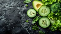 Fresh green vegetables including avocado, cucumber slices, broccoli and leafy greens arranged on dark slate background for healthy eating and nutrition.