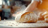 Fresh baked artisan bread loaf with flour dust cloud on wooden surface in warm bakery kitchen setting for culinary and food preparation concepts.