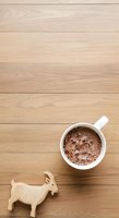 Overhead view of white mug filled with hot chocolate topped with cocoa powder on light wood surface, wooden goat figurine beside it, flat lay composition for beverage and lifestyle content.