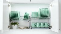 Organized kitchen cabinet with mint green dishes, plates, bowls, and mugs neatly stacked on a chrome drying rack with a small plant.