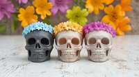 Decorative skulls with floral crowns arranged in row on marble surface, pink and orange flowers in background create festive Day of Dead atmosphere.