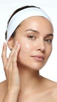 Young woman applying moisturizer cream to face during skincare routine wearing white headband for beauty and wellness treatment.