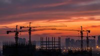 Construction site with tower cranes silhouetted against dramatic orange and purple sunset sky. Urban development and building industry at golden hour.