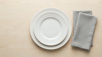 White ceramic plates stacked on beige wooden surface with gray linen napkin, flat lay composition for dining and tableware design projects.