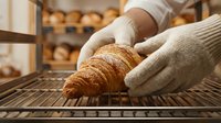 Baker in white gloves holding fresh croissant on cooling rack in bakery with blurred shelves of baked goods in background. Artisan bread production.