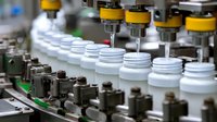 Pharmaceutical manufacturing line with automated filling machines inserting components into white plastic containers on conveyor belt in factory setting.