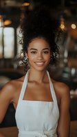 Young African American woman with natural curly hair wearing white apron smiling warmly at camera in restaurant setting with soft bokeh lighting background.