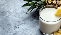 Fresh pineapple smoothie in glass with slice garnish and whole tropical fruit on gray concrete background, healthy summer drink with copy space.