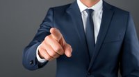 Businessman in navy blue suit pointing finger forward against gray background, conveying leadership and decision-making in professional setting.