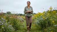 Middle-aged Caucasian woman botanist walking through wildflower meadow holding notebook, studying native plants in natural outdoor research setting.