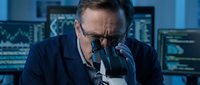 Middle-aged Caucasian male scientist in dark suit examining sample through microscope in modern laboratory with computer monitors displaying data charts.