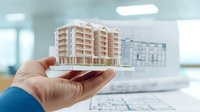 Architectural scale model of modern residential building held by architect's hand over blueprints in office, showing planning and development concept.