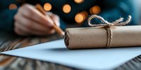 Rolled kraft paper scroll tied with rustic twine on blue surface, with person writing letter and warm bokeh lights in background, creating nostalgic atmosphere.