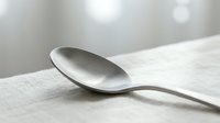Silver spoon resting on white linen fabric with soft bokeh background, suitable for food styling, culinary content, and tableware design projects.