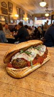 Meatball sandwich with melted cheese and marinara sauce on sesame seed roll, served in casual dining restaurant with warm lighting and blurred diners in background.