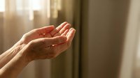 Elderly hands cupped together in prayer or meditation gesture with warm sunlight streaming through window creating peaceful spiritual moment.