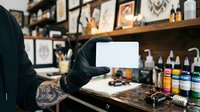 Tattoo artist with tattooed arm wearing black gloves holding blank white card in studio workspace with supplies, inks, and artwork on walls for design consultation.
