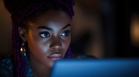 Young African woman with purple braids illuminated by blue light from screen, focused expression while working late at night on computer project.