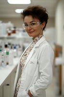 Young female scientist in laboratory wearing protective eyewear and white coat, with floral blouse and confident smile, standing near research equipment in modern facility.