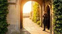Young woman in dark clothing standing in arched stone doorway with ivy-covered walls and sunlit pathway, suitable for travel, architecture, and lifestyle editorial use.