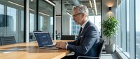 Mature Caucasian businessman in suit working on laptop at modern office desk near windows. Professional executive analyzing data in contemporary workplace.