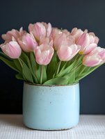 Fresh pink tulips arranged in rustic blue ceramic vase on white wooden surface against dark background for spring home decor and floral design projects.