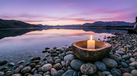 Glowing candle in stone bowl on pebble shore at twilight lake with mountain reflections and purple pink sky for meditation wellness spa relaxation concepts.
