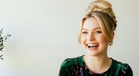 Young woman with blonde hair styled in an updo, fair complexion, and bright smile wearing a sequined green top against a neutral background.