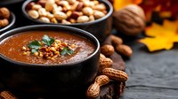 Homemade pumpkin soup garnished with parsley and nuts in black bowl, autumn setting with peanuts and walnuts on dark rustic background.