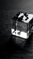Black and white artistic photo of transparent glass cube with reflections on dark textured surface creating dramatic shadows and light patterns.
