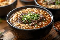 Spicy Asian noodle soup with tender braised beef, scallions and ramen in rich broth served in traditional black ceramic bowl on wooden table. Close up view.