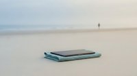 Smartphone with turquoise towel on sandy beach with ocean and distant person in background, suitable for travel, technology, and lifestyle marketing materials.