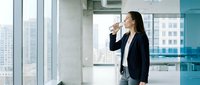 Professional Asian businesswoman drinking coffee in modern office with city view through large windows during morning break.