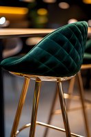 Luxurious emerald green velvet bar stool with quilted pattern and gold metal legs against blurred restaurant background, modern furniture design detail.