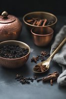 Moody dark composition with loose black tea leaves in copper bowl, star anise in golden spoon, cinnamon sticks and traditional teapot on dark background.