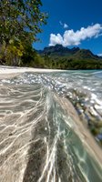 Crystal clear wave breaking on tropical beach with white sand, lush green mountains and vibrant blue sky. Split-level underwater photography captures pristine shoreline.