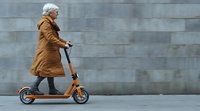 Senior Caucasian woman in brown winter coat riding orange electric scooter along grey wall, side view. Modern urban mobility for active aging lifestyle.