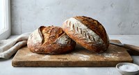 Freshly baked artisan sourdough bread loaves with golden crust on wooden cutting board near window, rustic bakery style for food photography and cooking content.