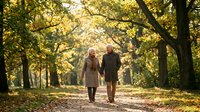 Senior couple walking together through autumn park with golden sunlight filtering through trees, enjoying peaceful moment in nature.