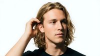 Young adult male with shoulder-length wavy blonde hair and fair complexion, wearing black casual clothing, touching his hair in thoughtful pose against white background.