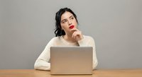 Young woman in beige sweater thinking while working on laptop at wooden desk, finger on lips in contemplative pose against neutral background.