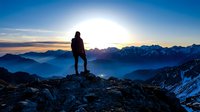 Silhouetted hiker stands on rocky mountain peak at sunrise, gazing at vast alpine landscape with layered mountain ranges and golden sky. Adventure and exploration.