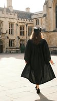 Female graduate in black cap and gown walking away from historic university building, celebrating academic achievement and educational milestone.