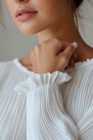 Close-up portrait of young Asian woman in white pleated blouse with ruffled collar touching neck, showing natural makeup and plump lips. Soft beauty concept.