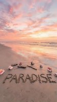 Word Paradise written in sand on a tropical beach at sunset with pink sky, seashells, and ocean waves in the background.