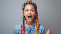 Young African American woman with colorful braids and denim shirt expressing extreme excitement and joy against gray background, showing genuine positive emotion.