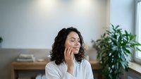 Thoughtful woman with curly dark hair wearing white shirt, resting chin on hand in modern office space with plants and natural light for business, corporate, and professional concepts.