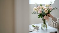 Woman arranging fresh pink and white roses with eucalyptus in glass vase on white table in bright modern interior for home decor and floral design.